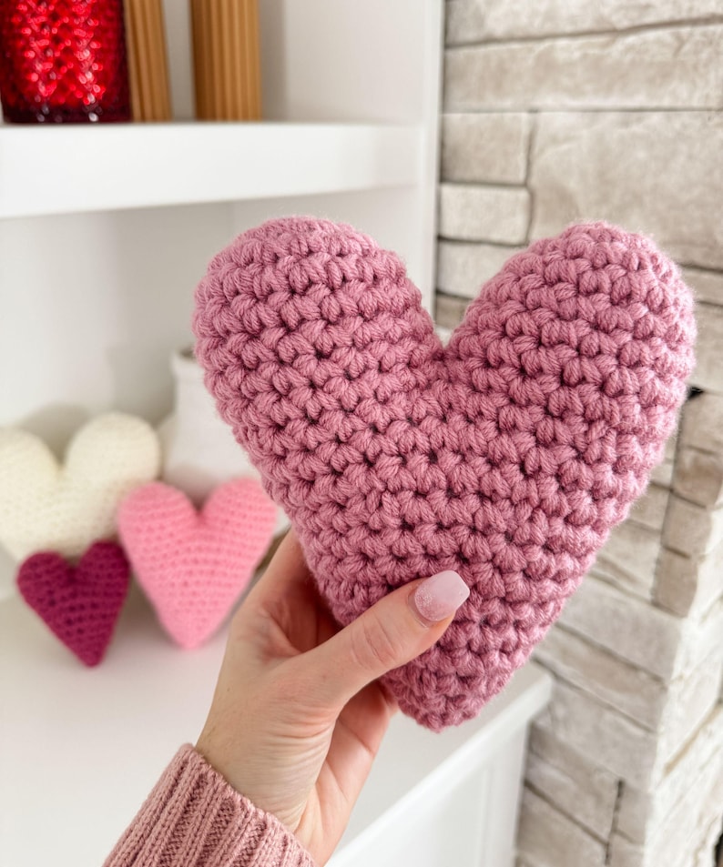 Pu&ograve; includere: Un cuscino a forma di cuore in crochet rosa. Il cuscino &egrave; tenuto in una mano.