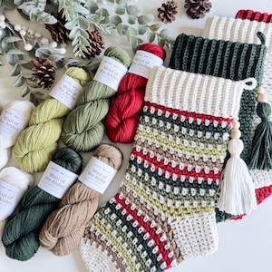 May include: A selection of Christmas stockings and yarn skeins in various colours. The stockings are hand-crocheted with white, green, red, and brown yarn. The yarn skeins are labelled "Wool of the Andes" and are in shades of white, green, and red.