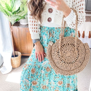 Puede incluir: Una mujer lleva un top blanco de crochet y una falda maxi floral azul. Lleva un bolso tote grande, redondo y de crochet marrón.