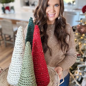 May include: Four crocheted Christmas trees in a white tray. The trees are white, green, and red. The trees are decorated with white and gold glitter.