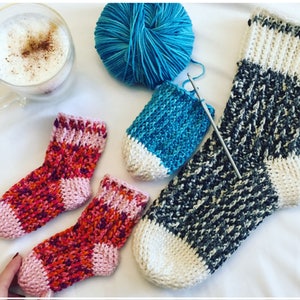 May include: Three pairs of crocheted socks in various colors, including pink, orange, blue, white, and gray. The socks are laid out on a white surface with a cup of coffee and a ball of yarn.