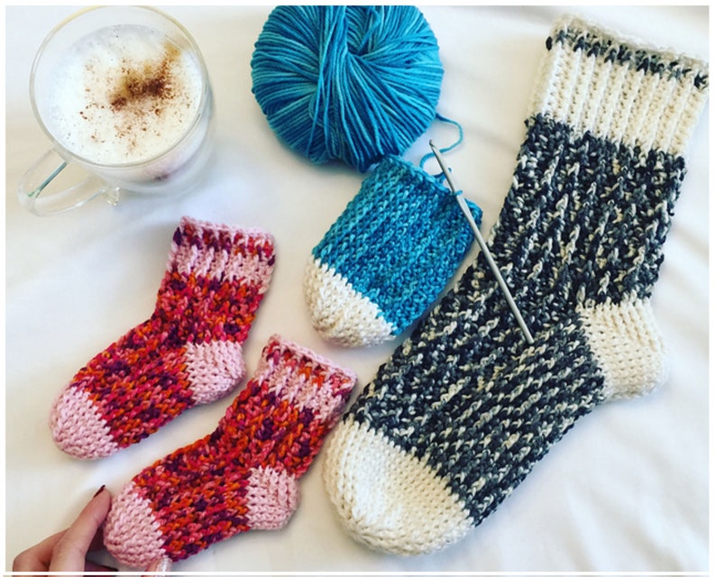 May include: Three pairs of crocheted socks in various colors, including pink, orange, blue, white, and gray. The socks are laid out on a white surface with a cup of coffee and a ball of yarn.