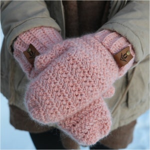 May include: A pair of pink crocheted mittens with brown leather accents. The mittens have a textured stitch pattern and are folded over at the wrist.