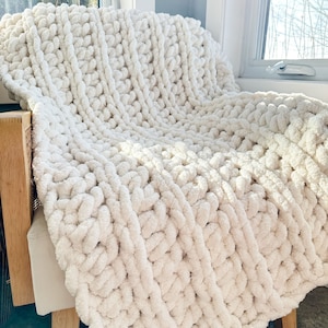 Peut inclure: Une couverture en tricot blanc &eacute;paisse avec une texture douce et moelleuse. La couverture est drap&eacute;e sur une chaise et a une ambiance d&eacute;tendue et confortable.
