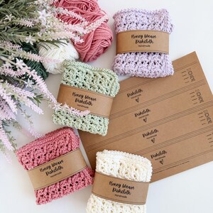 Puede incluir: Varios pa&ntilde;os de cocina Honey Weave hechos a mano en tonos rosa, verde, lavanda y crema. Cada pa&ntilde;o est&aacute; envuelto con una banda de papel marr&oacute;n que dice "Honey Weave Dishcloth Handmade". Tambi&eacute;n se ven ovillos de lana y elementos florales.