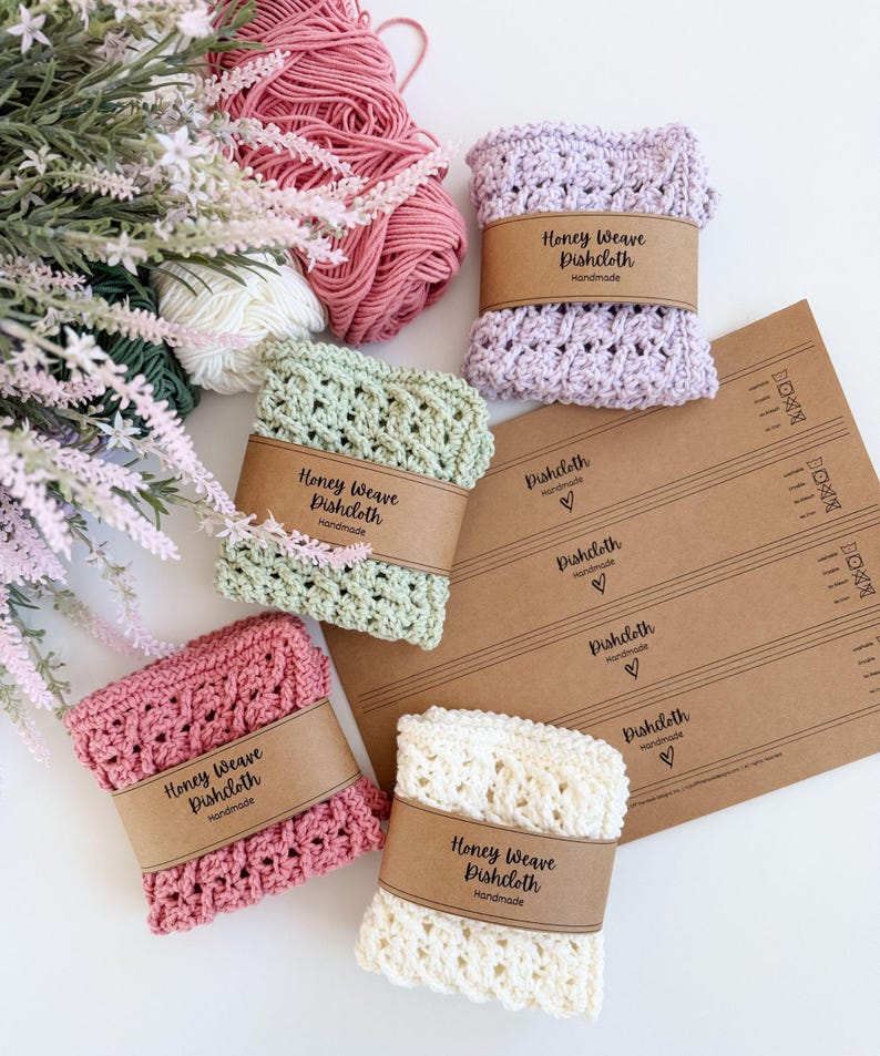 Puede incluir: Varios pa&ntilde;os de cocina Honey Weave hechos a mano en tonos rosa, verde, lavanda y crema. Cada pa&ntilde;o est&aacute; envuelto con una banda de papel marr&oacute;n que dice "Honey Weave Dishcloth Handmade". Tambi&eacute;n se ven ovillos de lana y elementos florales.