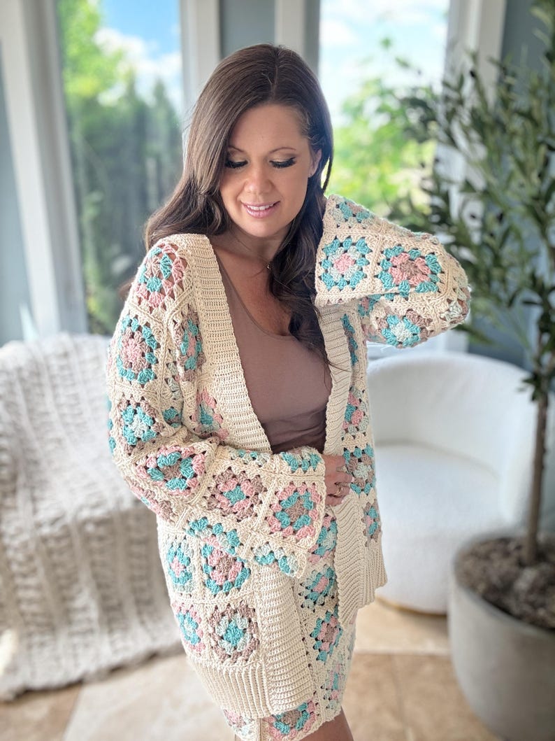 May include: A crocheted cardigan featuring a granny square design in cream, pink and blue hues. The cardigan has a long, open front and bell sleeves.
