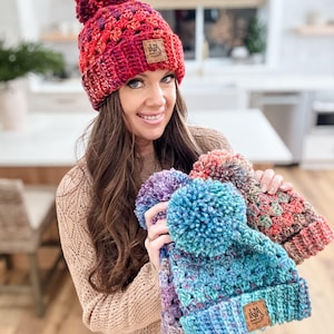 May include: Three crocheted hats with pom-poms in various colors, including red, blue, and purple. Each hat has a small brown leather tag with the text "M&M Handmade" on it.