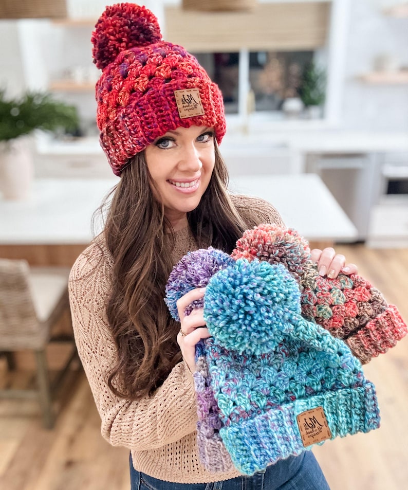 May include: Three crocheted hats with pom-poms in various colors, including red, blue, and purple. Each hat has a small brown leather tag with the text "M&M Handmade" on it.
