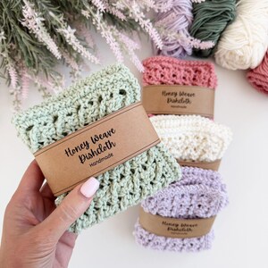 Puede incluir: Pa&ntilde;os de cocina Honey Weave hechos a mano en tonos pastel verde, rosa y lavanda. Cada pa&ntilde;o est&aacute; envuelto con una banda de papel marr&oacute;n que dice "Honey Weave Dishcloth Handmade". Madejas de hilo y acentos florales en el fondo.