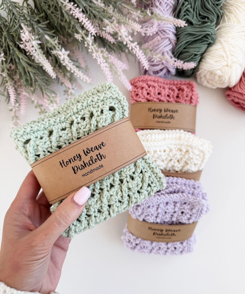 Puede incluir: Pa&ntilde;os de cocina Honey Weave hechos a mano en tonos pastel verde, rosa y lavanda. Cada pa&ntilde;o est&aacute; envuelto con una banda de papel marr&oacute;n que dice "Honey Weave Dishcloth Handmade". Madejas de hilo y acentos florales en el fondo.
