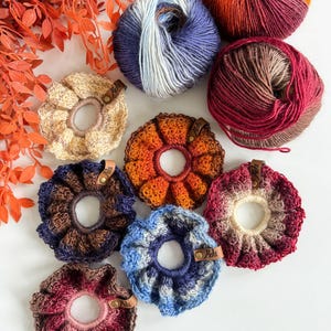 May include: A collection of handmade crochet scrunchies in various colors, including beige, orange, blue, and burgundy. Each scrunchie features a unique color pattern and is paired with a ball of yarn in coordinating shades. The scrunchies have small leather tags.