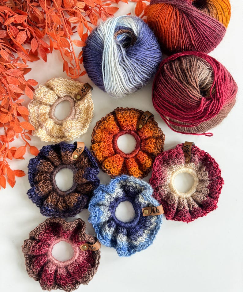 May include: A collection of handmade crochet scrunchies in various colors, including beige, orange, blue, and burgundy. Each scrunchie features a unique color pattern and is paired with a ball of yarn in coordinating shades. The scrunchies have small leather tags.