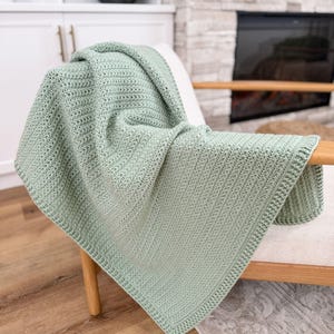 Puede incluir: Una manta de ganchillo color verde salvia que presenta un patr&oacute;n de puntos texturizados consistente en todo el cuerpo principal y un borde acanalado distintivo y ligeramente elevado que enmarca los bordes de la pieza textil rectangular.