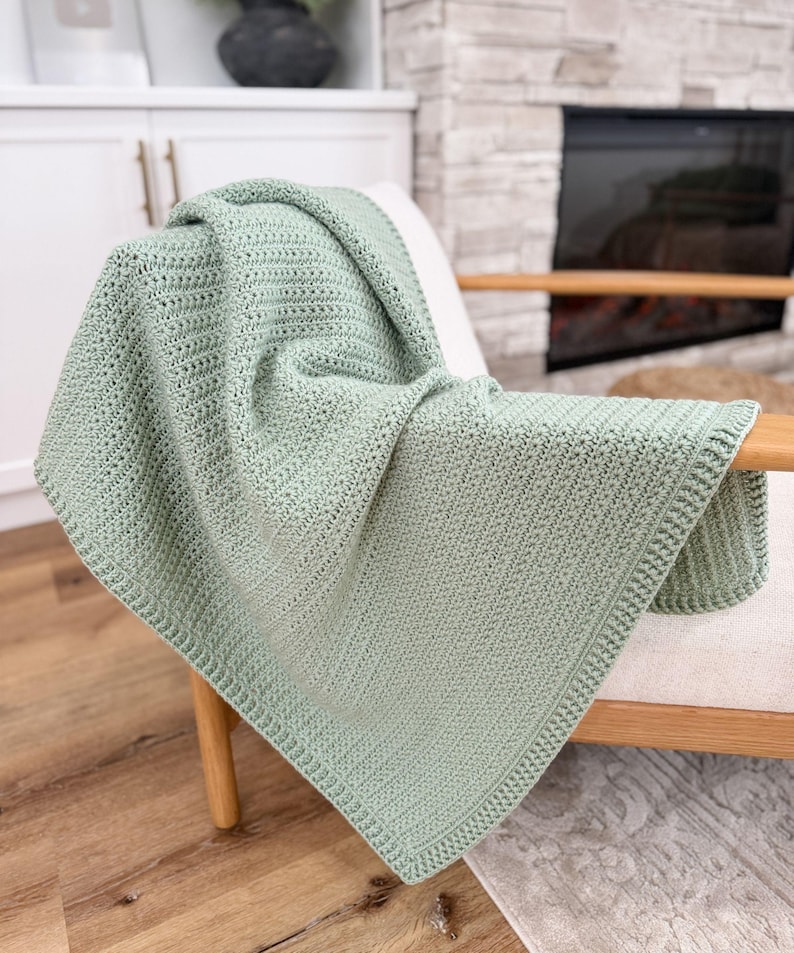 Puede incluir: Una manta de ganchillo color verde salvia que presenta un patr&oacute;n de puntos texturizados consistente en todo el cuerpo principal y un borde acanalado distintivo y ligeramente elevado que enmarca los bordes de la pieza textil rectangular.
