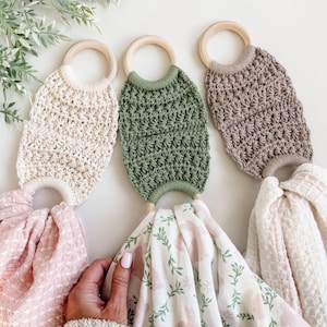 May include: Three macrame baby toys in cream, olive green, and taupe, each attached to a wooden ring. Below, soft pink and white patterned baby blankets are displayed. The blankets feature floral and bunny designs.
