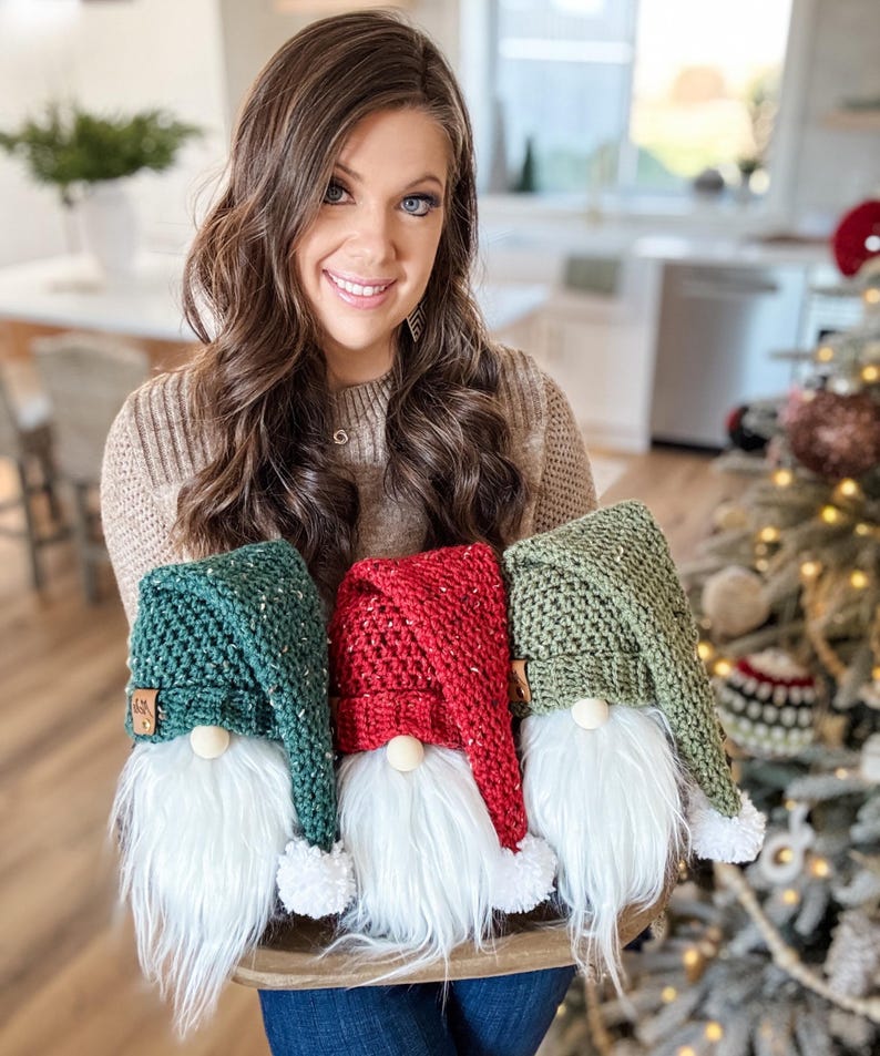 May include: Three crocheted gnomes with white fur beards. The gnomes are wearing green, red, and green hats with white pom-poms.