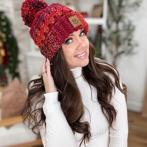 May include: A red, orange, and purple crocheted beanie with a pom pom on top. The beanie has a brown leather patch with the text "Ash & Arrow" on it.