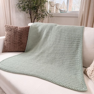 Puede incluir: Una manta de ganchillo color verde salvia que presenta una textura acanalada consistente en todo el cuerpo principal y un borde estructurado. La manta est&aacute; colocada sobre un sof&aacute; blanco junto a un coj&iacute;n marr&oacute;n con textura.