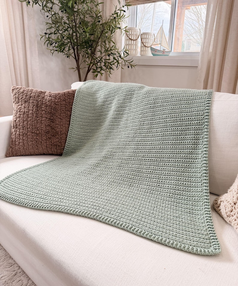 Puede incluir: Una manta de ganchillo color verde salvia que presenta una textura acanalada consistente en todo el cuerpo principal y un borde estructurado. La manta est&aacute; colocada sobre un sof&aacute; blanco junto a un coj&iacute;n marr&oacute;n con textura.
