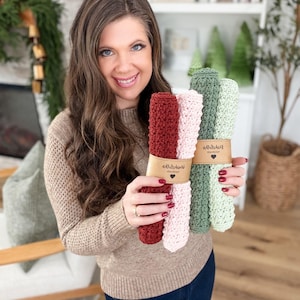 May include: A woman holds bundles of handmade crochet washcloths in shades of red, pink, and green. Each bundle is wrapped with a brown paper band. The washcloths are displayed in a well-lit room with a neutral background.