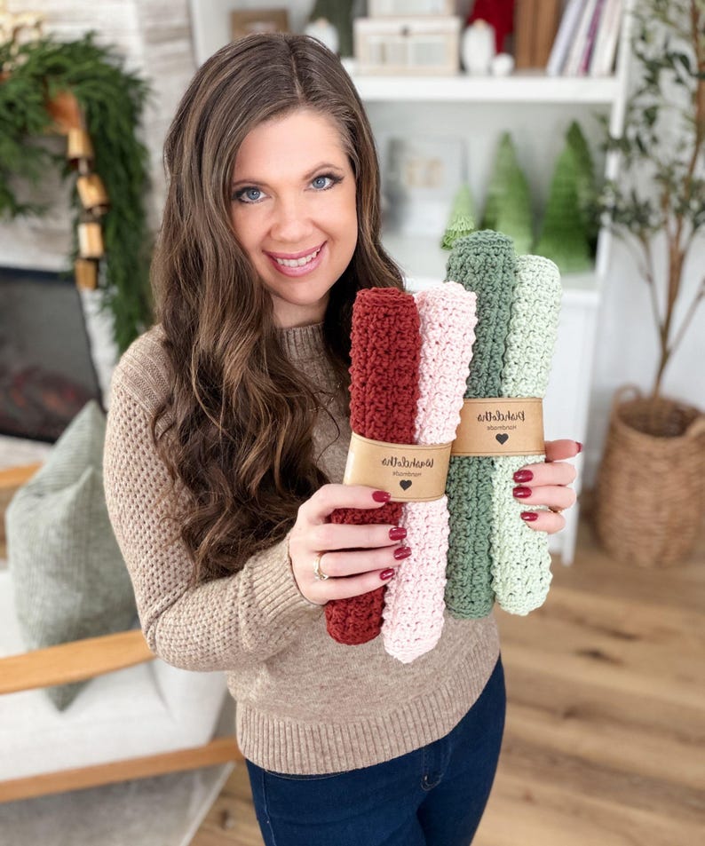 May include: A woman holds bundles of handmade crochet washcloths in shades of red, pink, and green. Each bundle is wrapped with a brown paper band. The washcloths are displayed in a well-lit room with a neutral background.