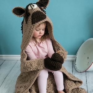 Pu&ograve; includere: Una coperta con cappuccio in crochet marrone con un design a testa di cavallo. La coperta &egrave; realizzata con filato marrone e bianco e presenta una testa di cavallo in crochet con occhi marroni e naso marrone. Un bambino indossa la coperta e siede su un pavimento di legno bianco.