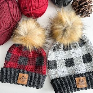 Op de afbeelding: Twee gebreide mutsen met nep bont pompoenen. De ene muts is rood en zwart geruit met een zwarte rand. De andere muts is wit en grijs geruit met een grijze rand. Beide mutsen hebben een bruin leren label met de tekst "Mels Off-Hook Designs".
