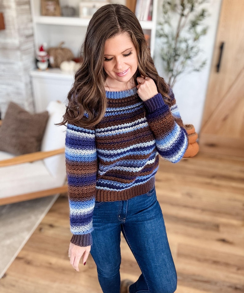 May include: A striped crochet sweater in shades of brown, blue, and white. The sweater has a crew neck and long sleeves. The model is wearing blue jeans.