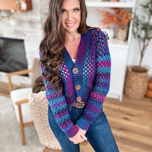 Op de afbeelding: Een kleurrijk gehaakt vest in paars, blauw en teal. De cropped trui heeft een V-hals, houten knopen en een textuursteekpatroon. De vrouw draagt het vest met een blauwe jeans en een bruine riem.