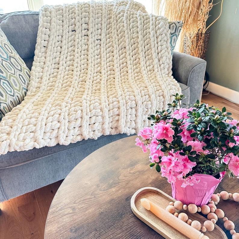 Peut inclure: Une couverture en tricot blanc &eacute;paisse drap&eacute; sur un canap&eacute; gris avec un arrangement de fleurs roses dans un pot sur une table en bois.