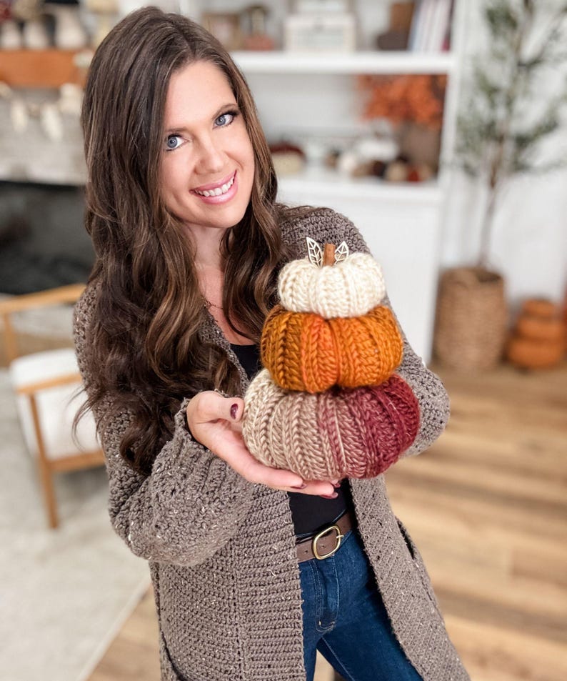K&ouml;nnte beinhalten: Ein Stapel von drei handgestrickten K&uuml;rbissen in Herbstfarben: Creme, Orange und ein Farbverlauf von Beige zu Burgunderrot. Jeder K&uuml;rbis hat einen Stiel und ist aufeinander gestapelt. Eine gem&uuml;tliche Herbstdekoration.