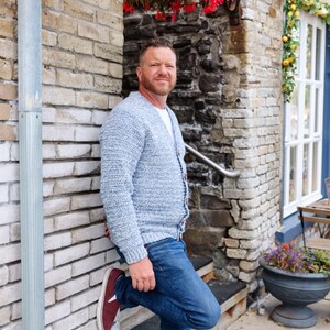 May include: A light blue knit cardigan with a button-up front. The cardigan is paired with blue jeans and burgundy sneakers. The person is leaning against a brick wall, with a stone wall and flowers in the background.