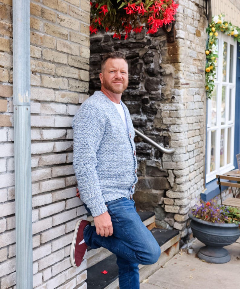 May include: A light blue knit cardigan with a button-up front. The cardigan is paired with blue jeans and burgundy sneakers. The person is leaning against a brick wall, with a stone wall and flowers in the background.