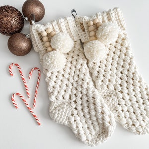 May include: A pair of white crocheted stockings with wooden beads and white pom-poms. The stockings are on a white surface.