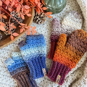 Peut inclure: Des mitaines sans doigts crochet&eacute;es aux couleurs automnales : bleu, marron et orange. Les gants, avec des poignets c&ocirc;tel&eacute;s, sont dispos&eacute;s sur une surface textur&eacute;e de couleur cr&egrave;me, avec une citrouille et des pommes de pin. Chaque gant a une petite &eacute;tiquette en cuir.