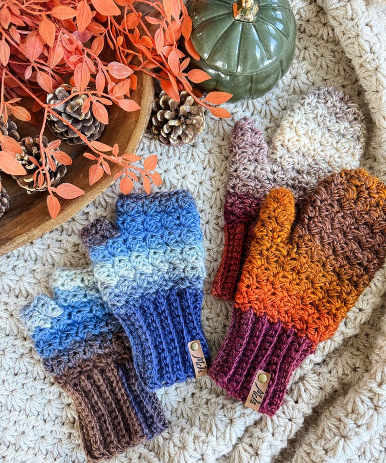 Peut inclure: Des mitaines sans doigts crochet&eacute;es aux couleurs automnales : bleu, marron et orange. Les gants, avec des poignets c&ocirc;tel&eacute;s, sont dispos&eacute;s sur une surface textur&eacute;e de couleur cr&egrave;me, avec une citrouille et des pommes de pin. Chaque gant a une petite &eacute;tiquette en cuir.