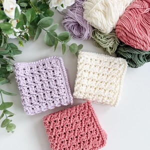 Puede incluir: Vista cenital de tres pa&ntilde;os de crochet en lavanda, crema y rosa, junto a madejas de hilo en tonos coordinados. Los pa&ntilde;os tienen un dise&ntilde;o de tejido abierto y texturizado. Vegetaci&oacute;n y flores blancas en el fondo.