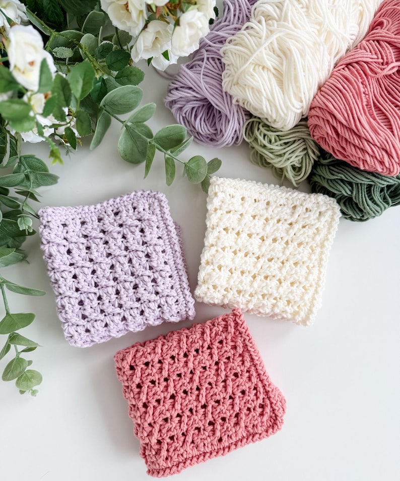 Puede incluir: Vista cenital de tres pa&ntilde;os de crochet en lavanda, crema y rosa, junto a madejas de hilo en tonos coordinados. Los pa&ntilde;os tienen un dise&ntilde;o de tejido abierto y texturizado. Vegetaci&oacute;n y flores blancas en el fondo.