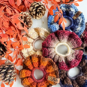 May include: A collection of handmade crochet scrunchies in various colors, including orange, cream, burgundy, and blue. The scrunchies are arranged with decorative orange leaves and pinecones, creating a fall-themed aesthetic. Each scrunchie has a small leather tag.