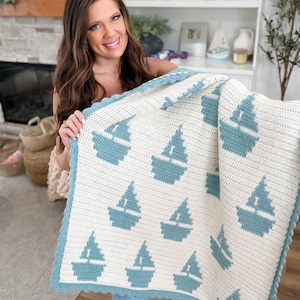 May include: A rectangular crochet blanket featuring a repeating pattern of teal sailboats on a cream-colored background. The blanket is finished with a scalloped teal border that frames the entire piece, showcasing a consistent stitch texture.