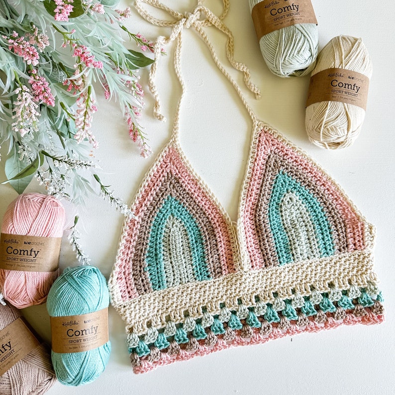 May include: A crocheted halter top with a rainbow pattern in shades of pink, green, and beige. The top has a scalloped hem and is made with a sport weight yarn.