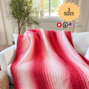 May include: A handmade, red and white ombre crochet blanket draped over a white sofa. The blanket features a textured stitch pattern and a gradient color scheme, transitioning from deep red to soft white. The image includes a text graphic that says "3 SIZES".