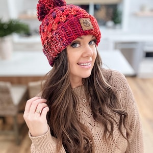 May include: A red and purple crocheted beanie with a pom pom on top. The beanie has a brown leather patch with the text "Ash & Co. Handmade" on it.