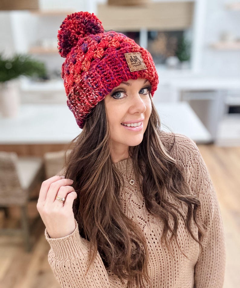 May include: A red and purple crocheted beanie with a pom pom on top. The beanie has a brown leather patch with the text "Ash & Co. Handmade" on it.