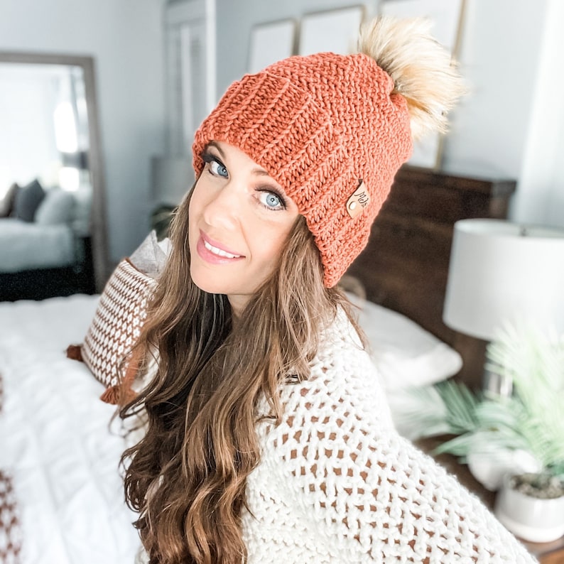 May include: A woman wearing a crocheted beanie with a faux fur pom pom in a burnt orange color. The beanie has a small leather tag with the word "Made" on it. She is wearing a white crocheted sweater.