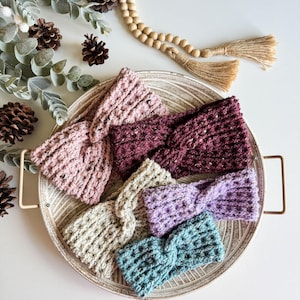 Puede incluir: Cinco diademas de crochet en una variedad de colores, incluyendo rosa, burdeos, beige, lavanda y azul verdoso, mostradas en una bandeja blanca con textura de madera.
