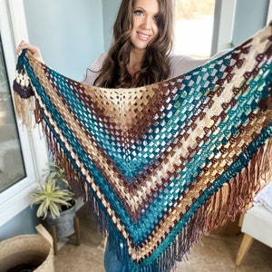 May include: A crocheted shawl with a brown, teal, and cream color pattern. The shawl has a fringe border and is held by a person.