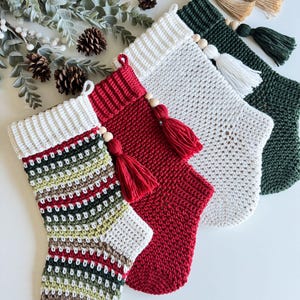 May include: Four handmade Christmas stockings in various colours. The stockings are crocheted with white, red, green, and patterned yarn. Each stocking has a white cuff, a wooden bead, and a tassel. The stockings are arranged on a white surface.