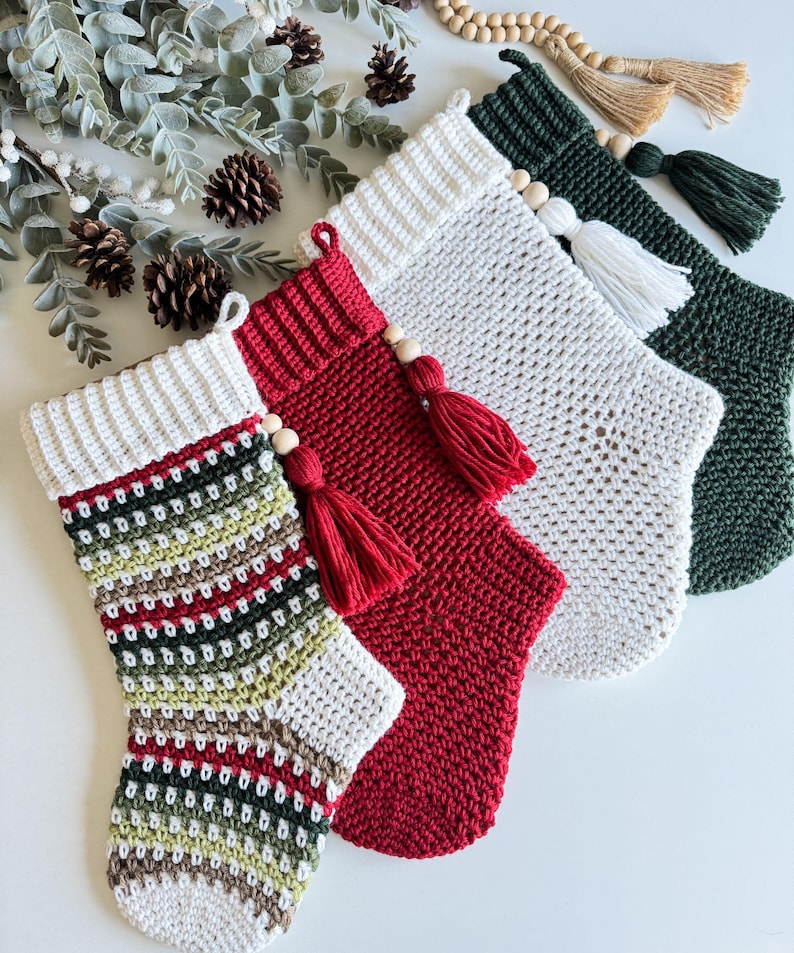 May include: Four handmade Christmas stockings in various colours. The stockings are crocheted with white, red, green, and patterned yarn. Each stocking has a white cuff, a wooden bead, and a tassel. The stockings are arranged on a white surface.
